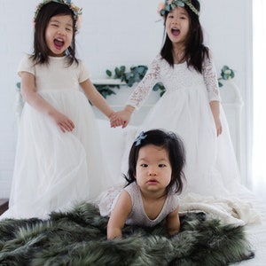 May include: Three young girls wearing white dresses and floral crowns. Two girls are standing and holding hands, while the third girl is sitting on a green faux fur blanket.