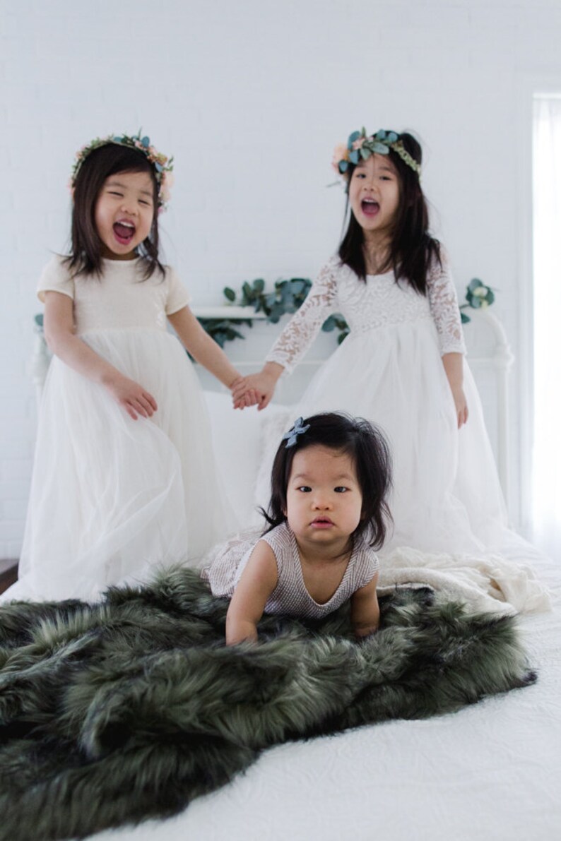 May include: Three young girls wearing white dresses and floral crowns. Two girls are standing and holding hands, while the third girl is sitting on a green faux fur blanket.