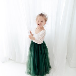 Pu&ograve; includere: Un abito lungo fino ai piedi con corpetto in pizzo bianco a maniche lunghe e scollo rotondo, abbinato a una voluminosa gonna in tulle color verde bosco scuro. Il capo presenta una parte superiore aderente e una gonna ampia e fluida.