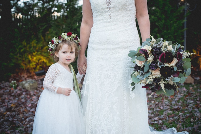 May include: A young girl in a white dress and flower crown holds hands with a bride in a white lace wedding dress. The bride is holding a bouquet of burgundy, white, and green flowers.