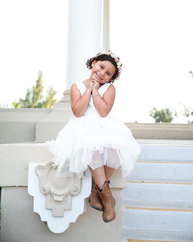 Op de afbeelding: Een jong meisje in een witte jurk en bruine laarzen zit op een witte trede met een bloemenkrans in haar haar. Ze glimlacht en kijkt naar de camera.