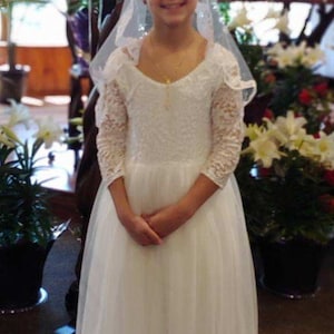 May include: A young girl in a white lace dress with a veil, standing in a church setting. She is smiling and holding her hands together in front of her.