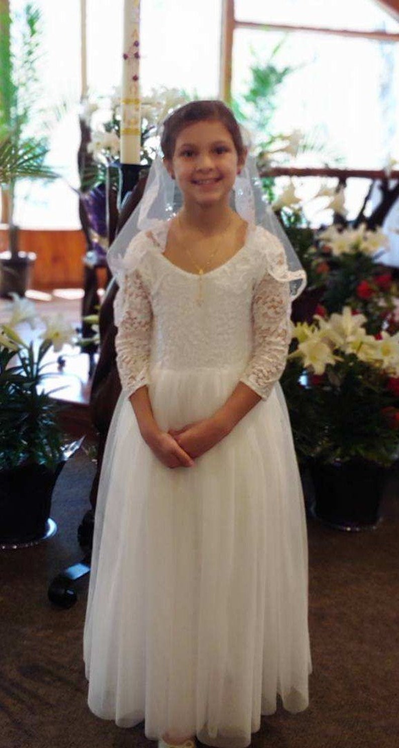 May include: A young girl in a white lace dress with a veil, standing in a church setting. She is smiling and holding her hands together in front of her.