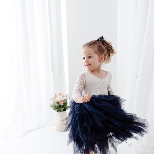 May include: A dress featuring a white lace bodice with three-quarter length sleeves and a voluminous, layered navy blue tulle skirt. The garment has a scoop neckline and a fitted waist, with the skirt flaring out in a tiered, ruffled design.