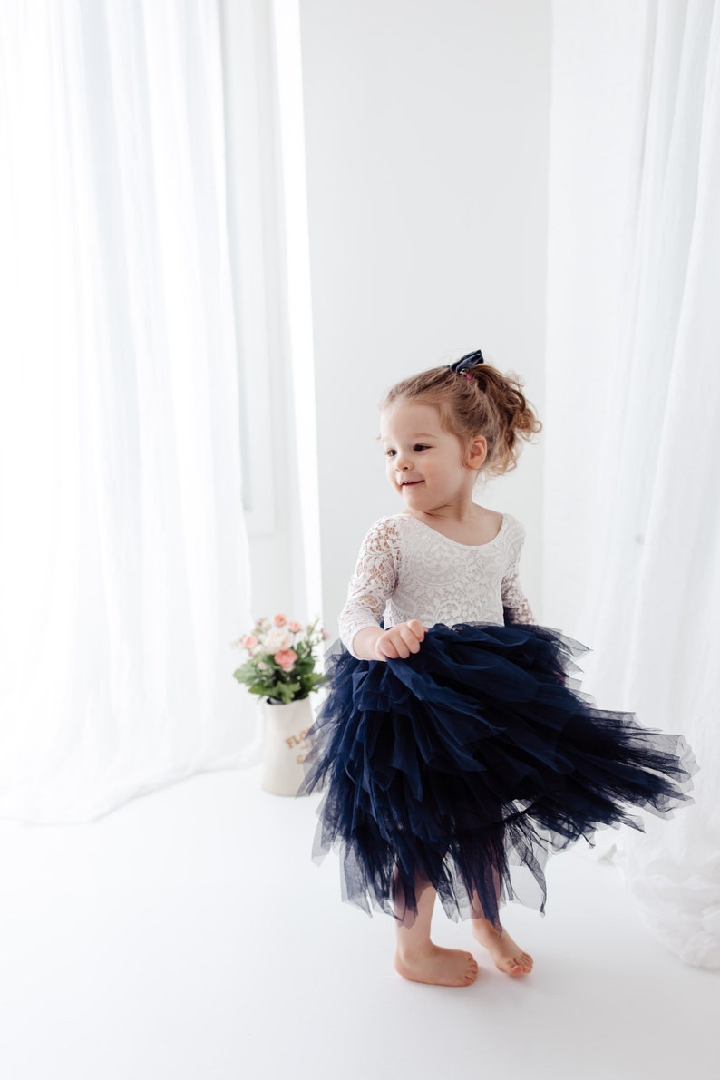 May include: A dress featuring a white lace bodice with three-quarter length sleeves and a voluminous, layered navy blue tulle skirt. The garment has a scoop neckline and a fitted waist, with the skirt flaring out in a tiered, ruffled design.