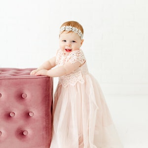 May include: A pale pink dress with a lace bodice and a layered tulle skirt, paired with a white floral headband. The dress features short lace sleeves and a scalloped neckline, creating a soft, layered appearance for the garment.