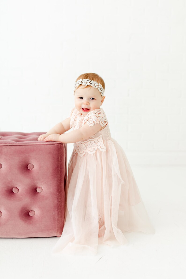 May include: A pale pink dress with a lace bodice and a layered tulle skirt, paired with a white floral headband. The dress features short lace sleeves and a scalloped neckline, creating a soft, layered appearance for the garment.