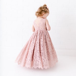 Pu&ograve; includere: Una bambina che indossa un abito di pizzo rosa chiaro con un orlo smerlato. L'abito &egrave; senza maniche e ha una scollatura rotonda.