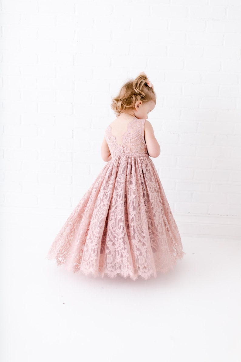 Pu&ograve; includere: Una bambina che indossa un abito di pizzo rosa chiaro con un orlo smerlato. L'abito &egrave; senza maniche e ha una scollatura rotonda.