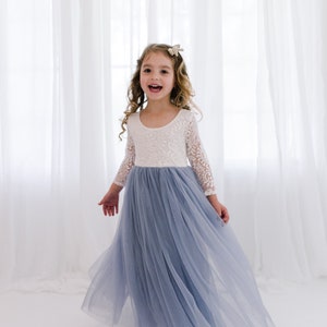 May include: A young girl wearing a white lace long-sleeved top and a light blue tulle skirt. She is smiling and twirling in front of a white curtain.