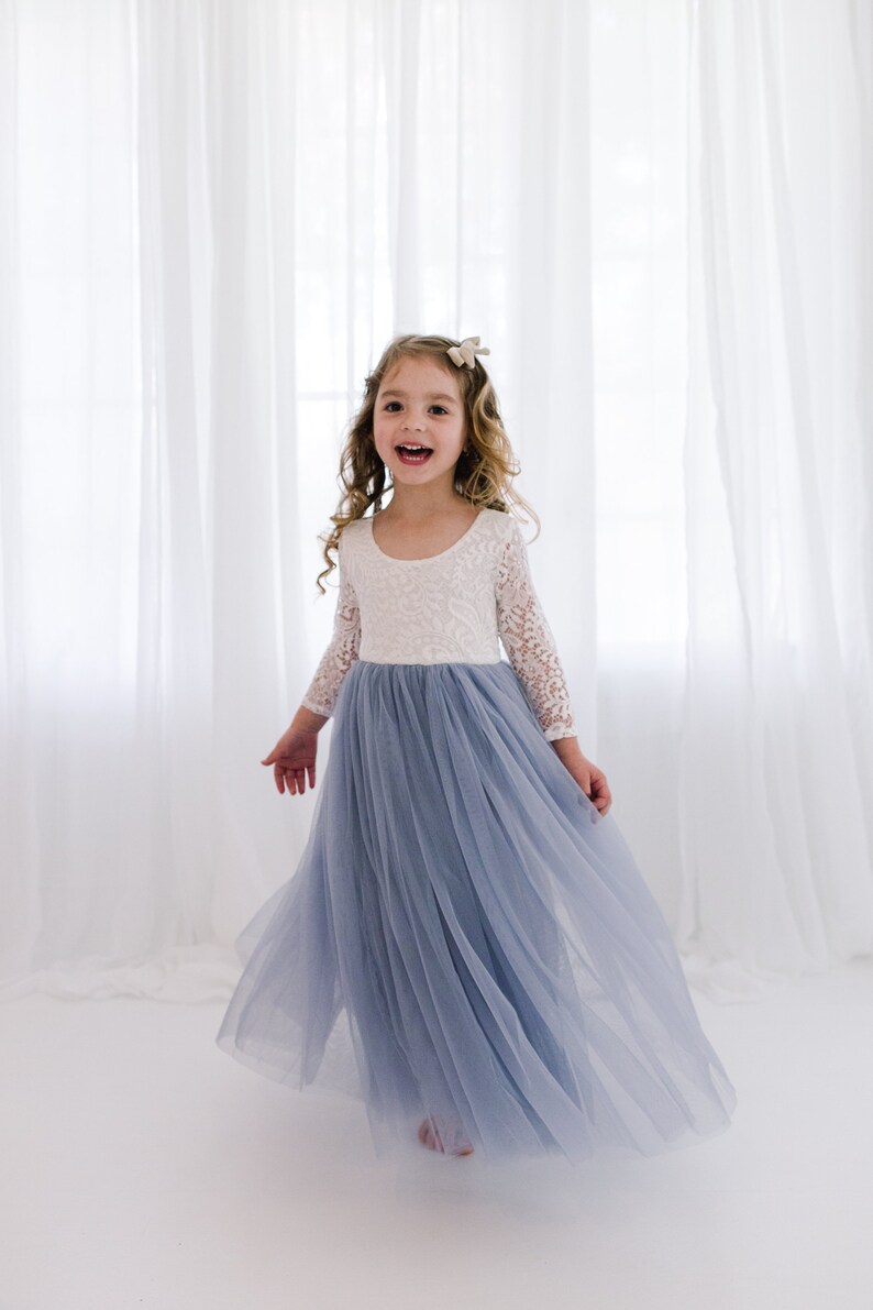 May include: A young girl wearing a white lace long-sleeved top and a light blue tulle skirt. She is smiling and twirling in front of a white curtain.