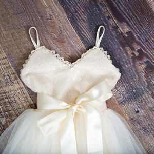 May include: A white tulle dress with a sequined bodice and a satin bow. The dress has a sweetheart neckline and thin straps.