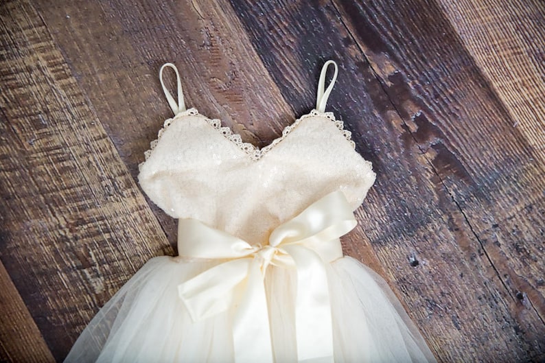 May include: A white tulle dress with a sequined bodice and a satin bow. The dress has a sweetheart neckline and thin straps.