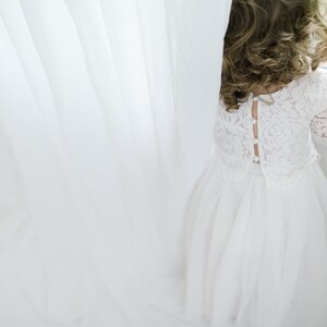 May include: A young girl in a white lace dress with a button-up back stands by a window, looking out.