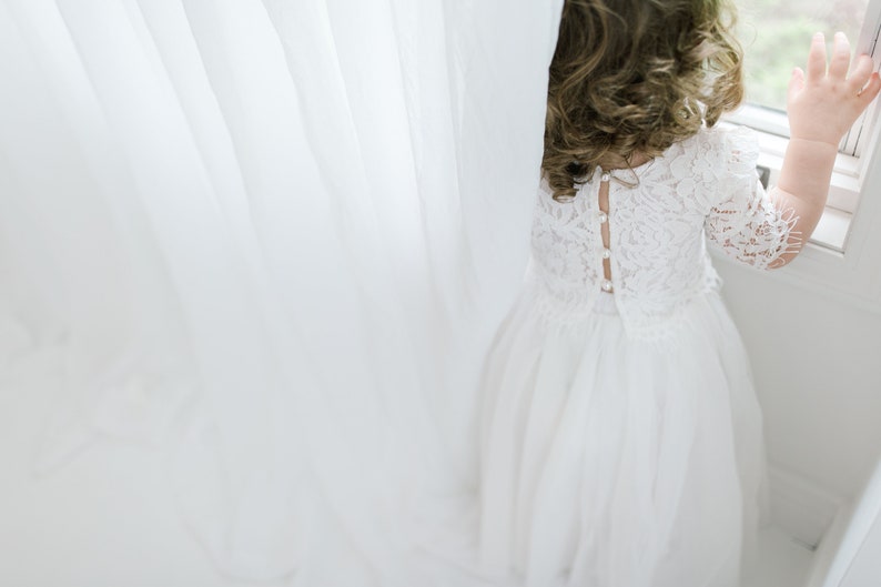 May include: A young girl in a white lace dress with a button-up back stands by a window, looking out.