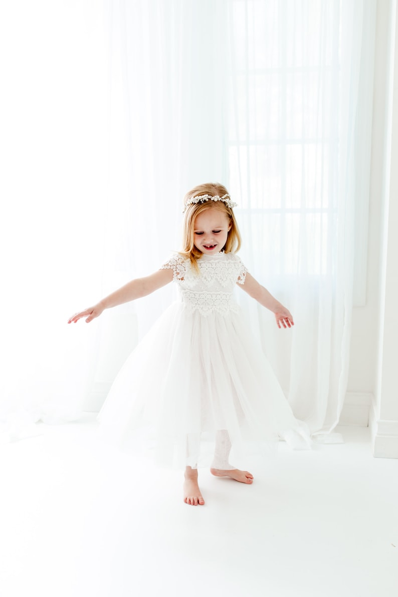 May include: A young girl wearing a white tulle dress with lace detailing and a flower crown. She is standing on a white background and smiling.