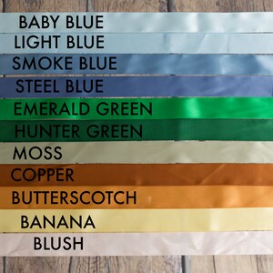 May include: A collection of eleven horizontal satin ribbons in various colors, each labeled with black text: Baby Blue, Light Blue, Smoke Blue, Steel Blue, Emerald Green, Hunter Green, Moss, Copper, Butterscotch, Banana, and Blush.