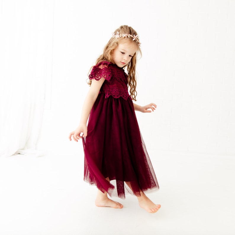May include: A young girl wearing a burgundy tulle dress with lace detailing and a floral crown. The dress has a full skirt and short sleeves.