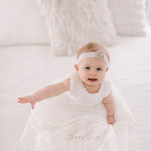 May include: A baby girl wearing a white lace dress and a headband with a jeweled embellishment sits on a white bed.