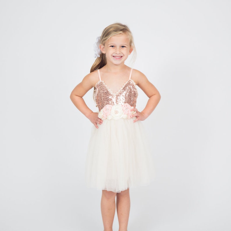 May include: A young girl wearing a white tulle dress with a rose gold sequin bodice and a pink flower sash. She is smiling and has her hands on her hips.