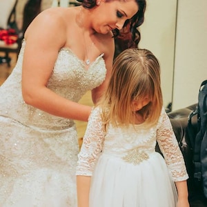 Puede incluir: Una novia con un vestido de novia blanco con cuentas ayuda a una ni&ntilde;a a ajustar su vestido blanco de encaje con una faja adornada con joyas.