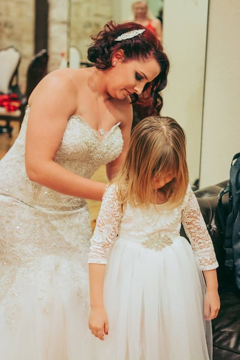 Puede incluir: Una novia con un vestido de novia blanco con cuentas ayuda a una ni&ntilde;a a ajustar su vestido blanco de encaje con una faja adornada con joyas.