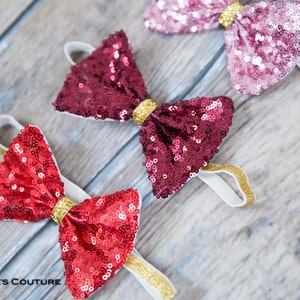May include: Three sequined bow headbands in red, burgundy, and pink with gold trim. The headbands are on a wooden surface.