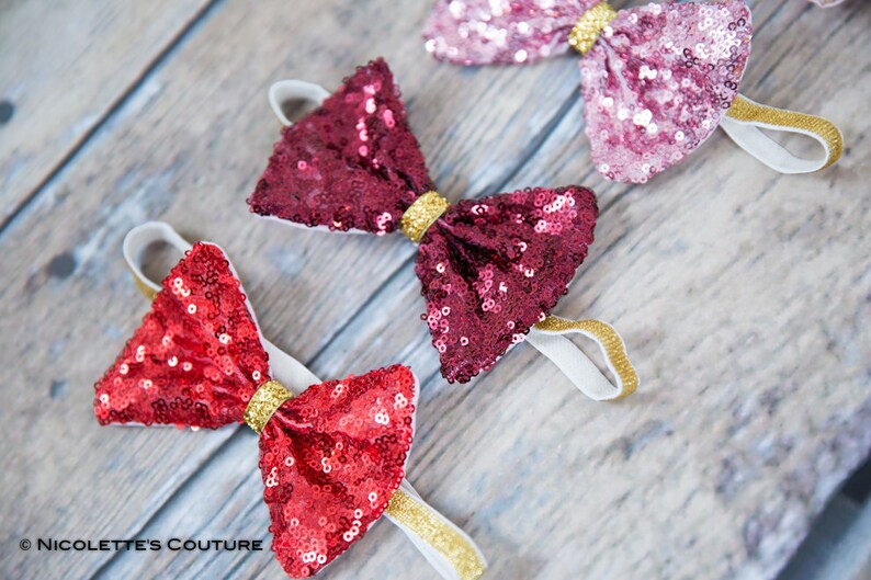 May include: Three sequined bow headbands in red, burgundy, and pink with gold trim. The headbands are on a wooden surface.