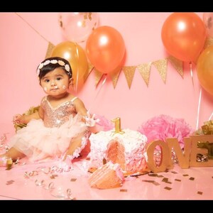 May include: A baby girl wearing a pink tutu dress and a flower crown sits in front of a pink cake with the number "1" on top. The cake is surrounded by pink and gold decorations, including balloons, confetti, and the word "LOVE" spelled out in gold letters.
