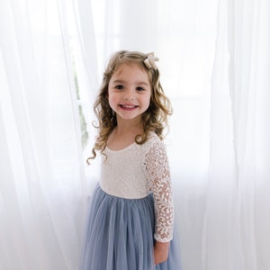 May include: A young girl with long curly brown hair wearing a white lace long-sleeved top and a light blue tulle skirt. She is smiling and looking at the camera.