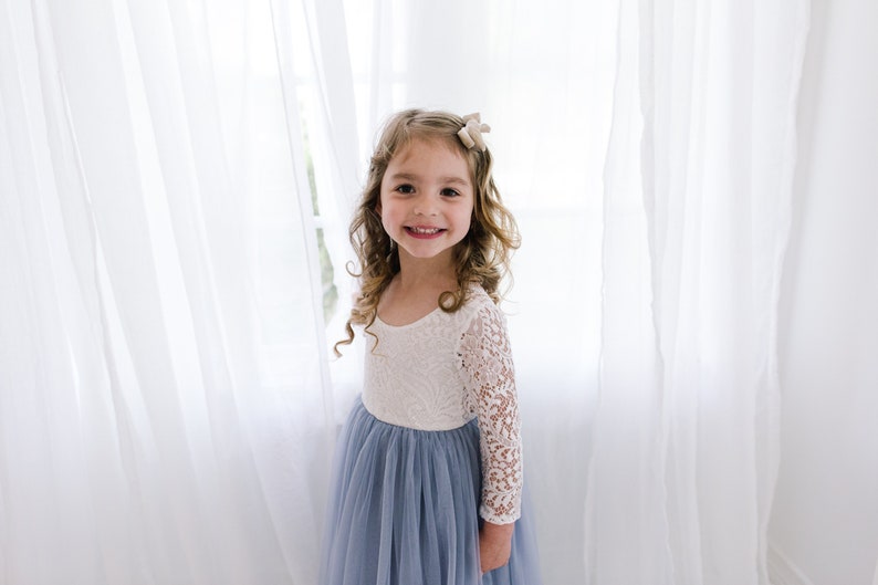 May include: A young girl with long curly brown hair wearing a white lace long-sleeved top and a light blue tulle skirt. She is smiling and looking at the camera.