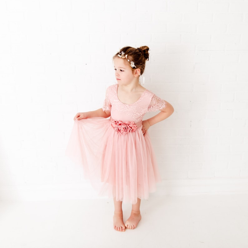Pode incluir: Uma menina pequena vestindo um vestido de tule rosa com detalhes em renda e uma faixa de flores. Ela est&aacute; usando uma faixa de cabe&ccedil;a dourada com flores brancas.