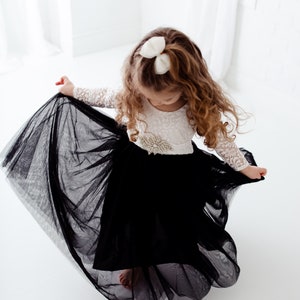 May include: A dress featuring a white lace long-sleeve bodice with a jeweled waist accent and a voluminous black tulle skirt. The garment includes a matching white bow hair accessory, creating a contrast between the delicate lace and the dark, layered tulle fabric that flares outward during movement.