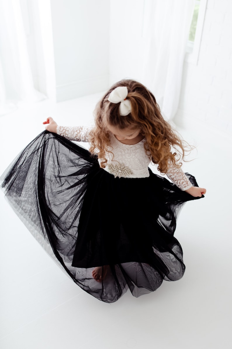 May include: A dress featuring a white lace long-sleeve bodice with a jeweled waist accent and a voluminous black tulle skirt. The garment includes a matching white bow hair accessory, creating a contrast between the delicate lace and the dark, layered tulle fabric that flares outward during movement.