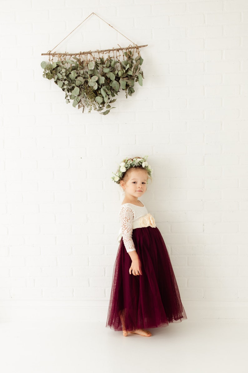 May include: A young girl wearing a white lace top with a burgundy tulle skirt and a flower crown. She is standing in front of a white brick wall with a greenery garland hanging above her.
