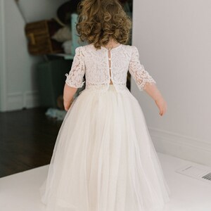 May include: A young girl wearing a white lace top with short sleeves and a long, flowing tulle skirt. The back of the top features button closures. The dress is a light ivory color, suitable for a special occasion.