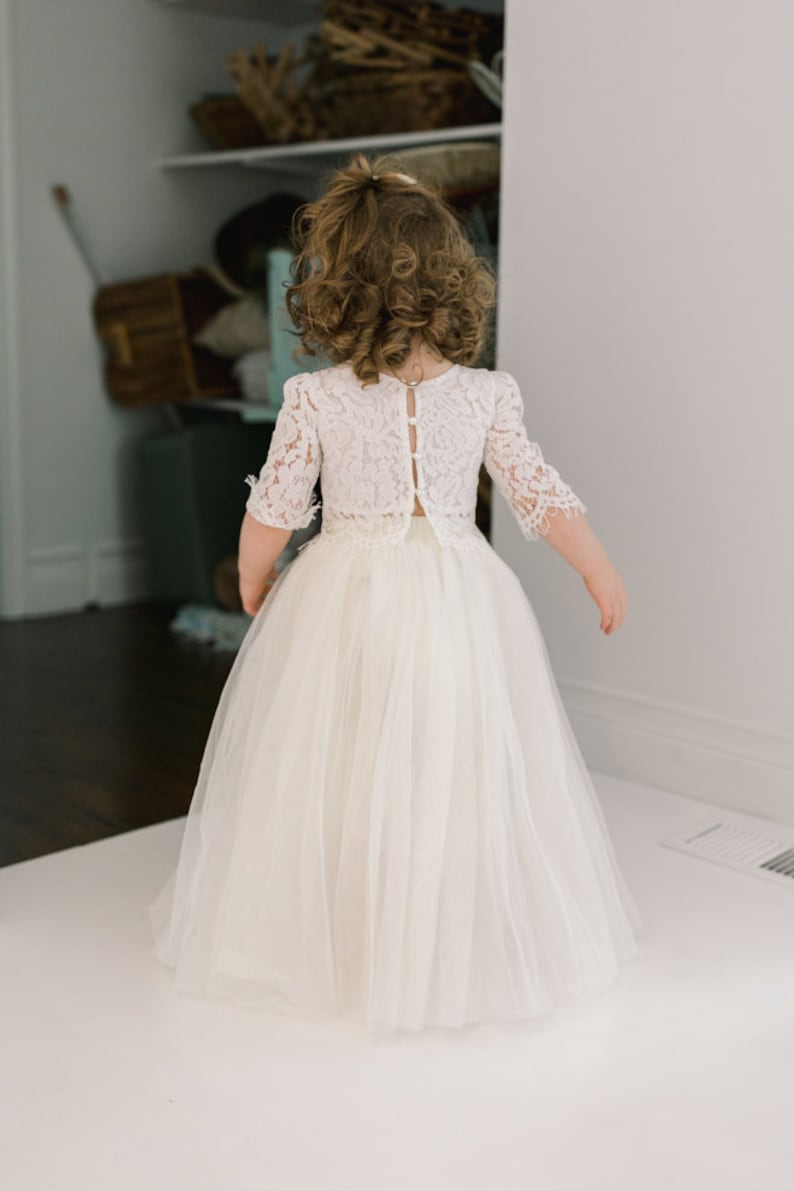May include: A young girl wearing a white lace top with short sleeves and a long, flowing tulle skirt. The back of the top features button closures. The dress is a light ivory color, suitable for a special occasion.
