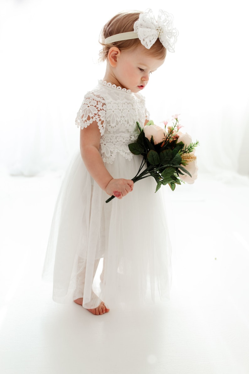 May include: A child in a white lace dress and a matching headband with a large bow. The child holds a bouquet of pink and cream flowers with green leaves. The dress has a tulle skirt, creating a soft, flowing look.