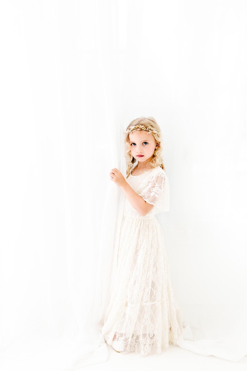 以下が含まれることがあります： 白い背景の前に立つ、白いレースのドレスと花冠をつけた若い女の子