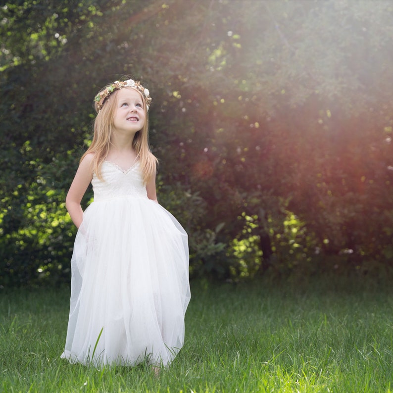 May include: A young girl wearing a white tulle dress with a floral crown, standing in a grassy field with a blurred background of trees.