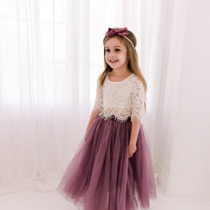May include: A young girl wearing a white lace crop top and a long, flowing mauve tulle skirt. She is smiling and has a mauve velvet bow headband in her hair.