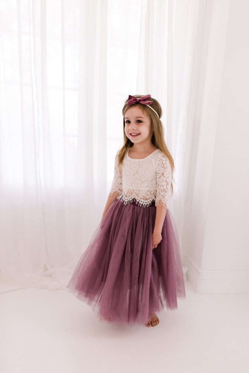 May include: A young girl wearing a white lace crop top and a long, flowing mauve tulle skirt. She is smiling and has a mauve velvet bow headband in her hair.