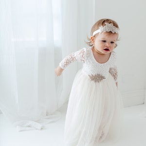 Pu&ograve; includere: Una bambina che indossa un abito bianco in pizzo con gonna di tulle e una fascia scintillante. L'abito ha una cintura con una fibbia gioiello.