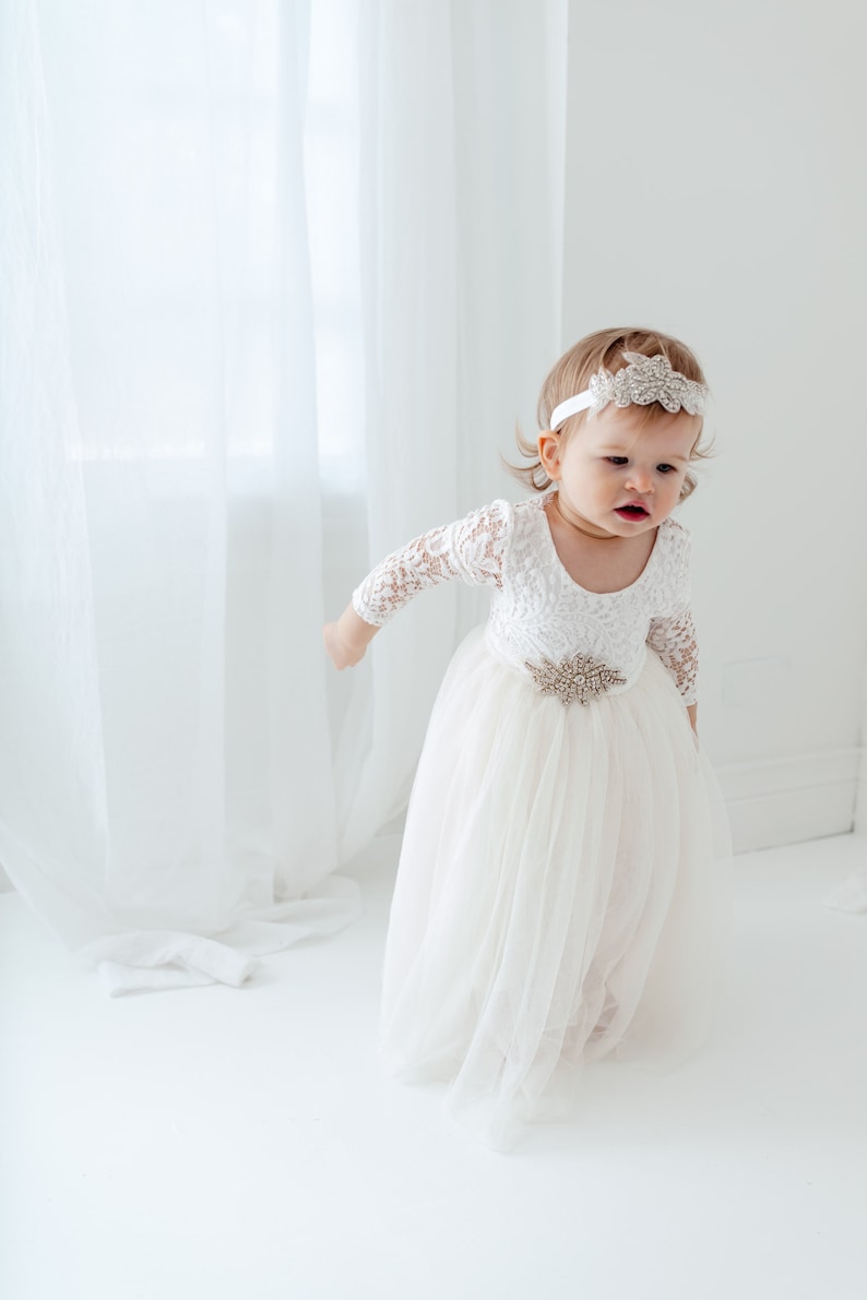 Pu&ograve; includere: Una bambina che indossa un abito bianco in pizzo con gonna di tulle e una fascia scintillante. L'abito ha una cintura con una fibbia gioiello.