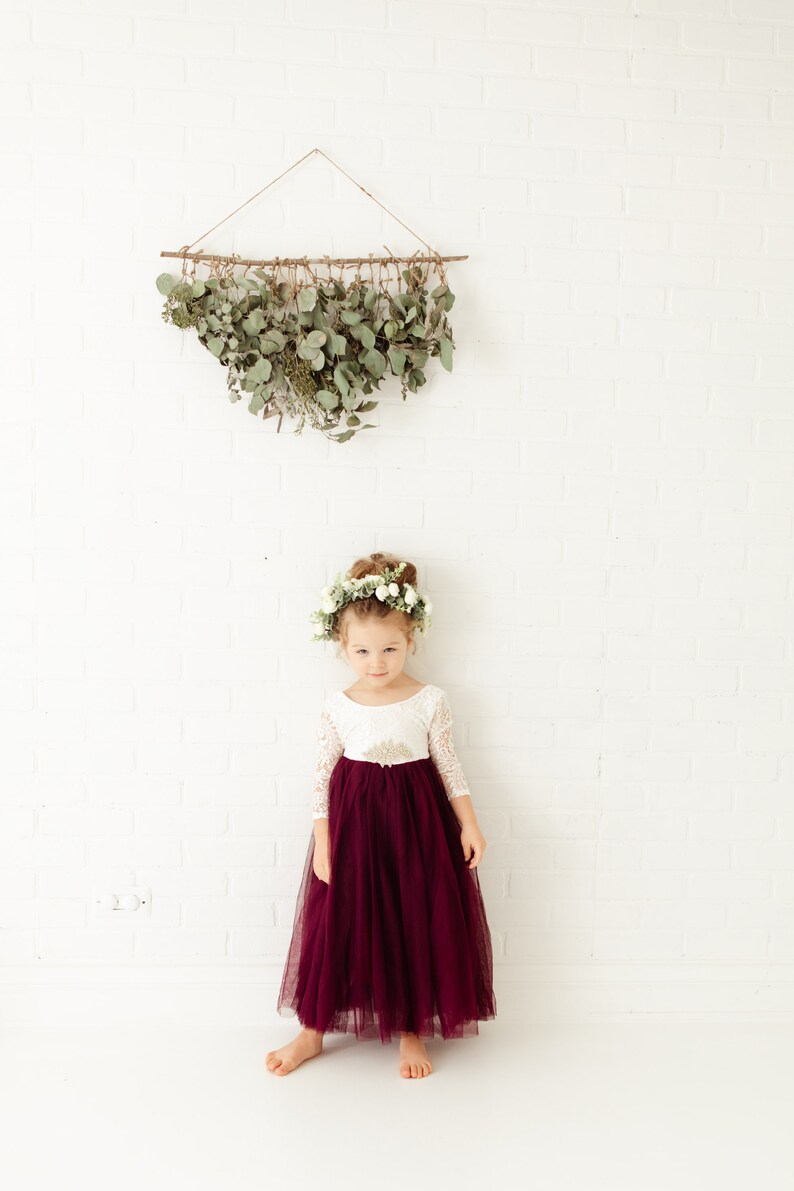 May include: A floor-length dress featuring a white lace bodice with three-quarter length sleeves and a deep burgundy tulle skirt. The waist is accented with a gold-toned embellishment. A floral crown with white flowers sits on the head.