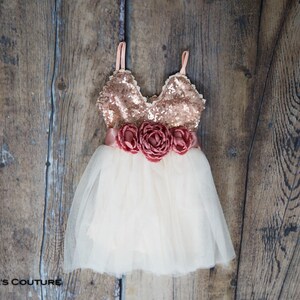 May include: A white tulle dress with a rose gold sequin bodice and a matching rose gold satin sash with three large rose-coloured fabric flowers.