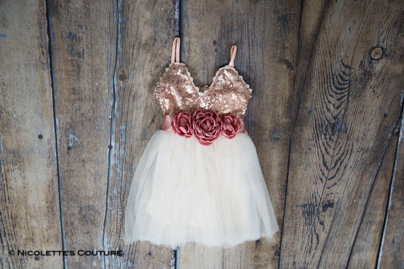 May include: A white tulle dress with a rose gold sequin bodice and a matching rose gold satin sash with three large rose-coloured fabric flowers.