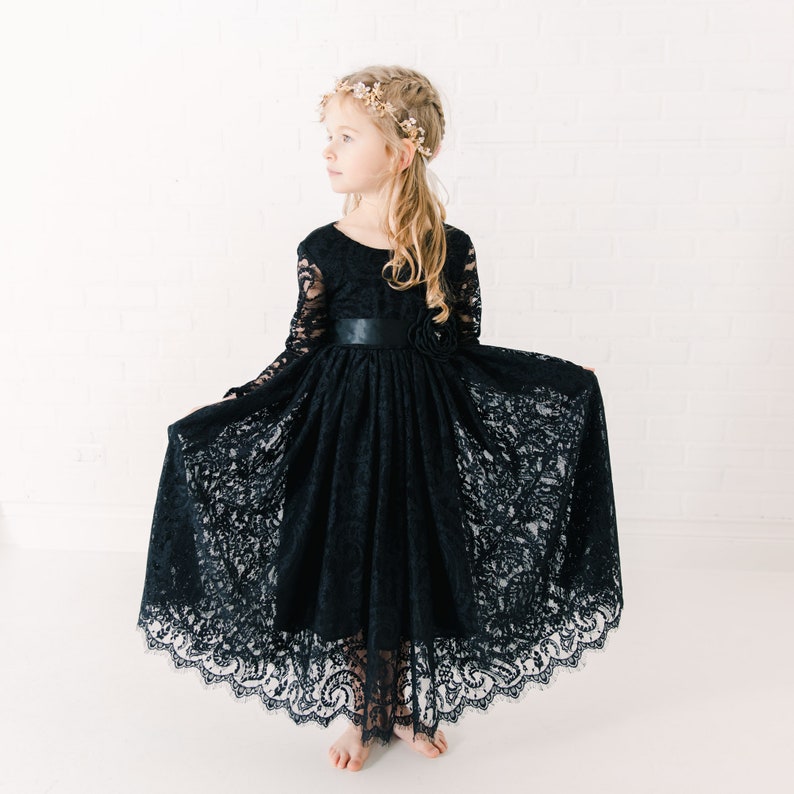 May include: A floor-length black lace dress featuring long sleeves, a round neckline, and a solid black satin waistband with a decorative fabric flower accent. The skirt is full and voluminous, showcasing an intricate floral lace pattern throughout.