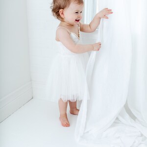 Puede incluir: Una ni&ntilde;a peque&ntilde;a con un vestido blanco de tul est&aacute; de pie cerca de una ventana, sosteniendo una cortina blanca y mirando hacia un lado. La ni&ntilde;a sonr&iacute;e.