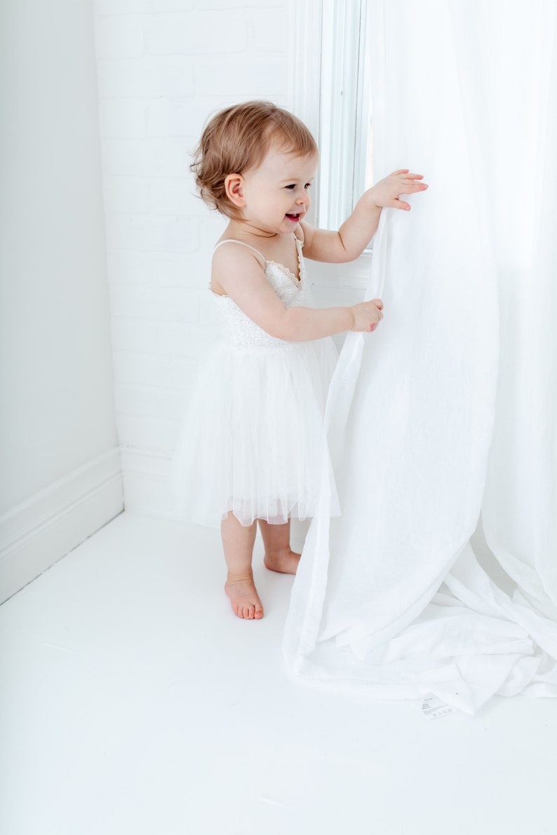 Puede incluir: Una ni&ntilde;a peque&ntilde;a con un vestido blanco de tul est&aacute; de pie cerca de una ventana, sosteniendo una cortina blanca y mirando hacia un lado. La ni&ntilde;a sonr&iacute;e.